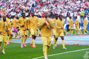 Soccer players in bright yellow uniforms walk onto a green field as fans cheer in the stands, with the central player adjusting a scarf around his neck.