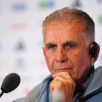 Man wearing a headset at a microphone during a press briefing, with a thoughtful expression on his face.”,