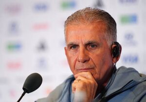Man wearing a headset at a microphone during a press briefing, with a thoughtful expression on his face.”,