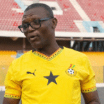 Man in a yellow Ghana football jersey with a black star and logo, wearing glasses, speaks on a stadium field.