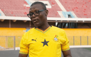Man in a yellow Ghana football jersey with a black star and logo, wearing glasses, speaks on a stadium field.