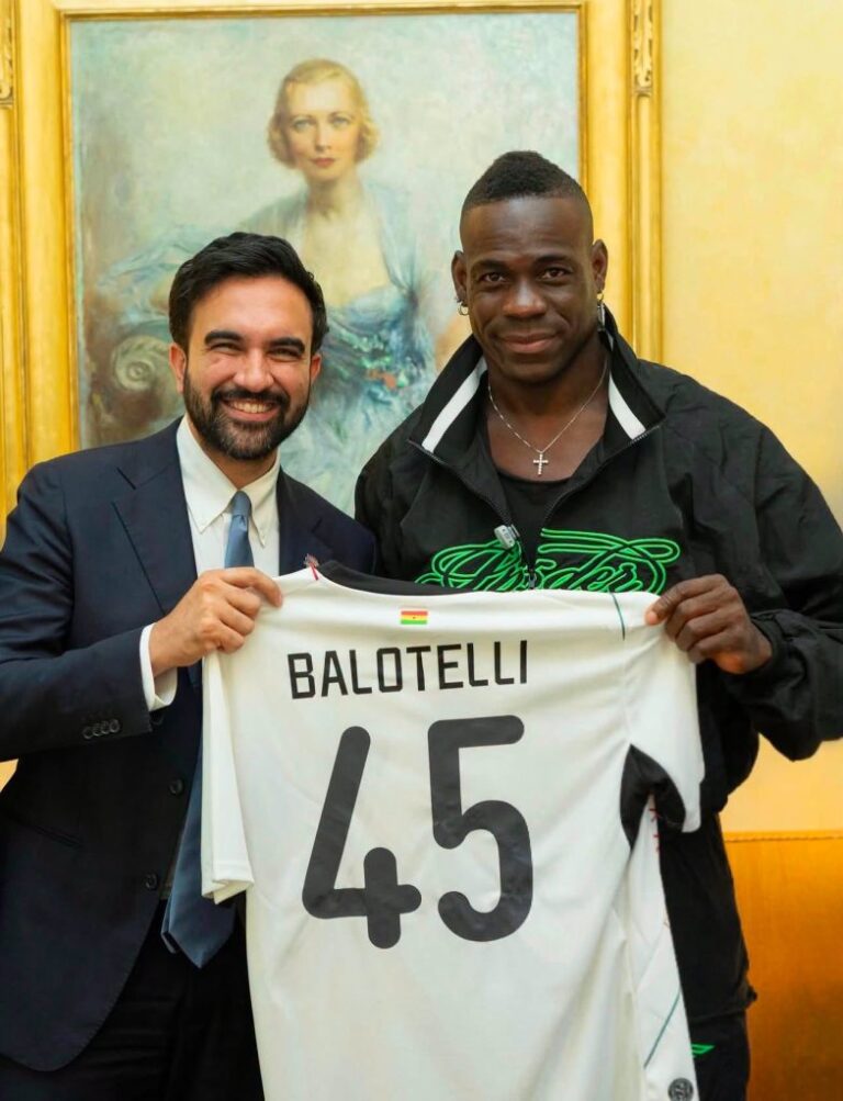Two men smile while holding a white soccer jersey that reads BALOTELLI 45, with a small flag, in front of a gold-framed painting.