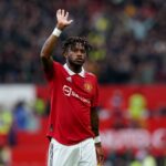 Manchester United footballer in a red kit raises his hand to the crowd during a match, stadium blurred in background.