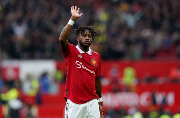 Manchester United footballer in a red kit raises his hand to the crowd during a match, stadium blurred in background.