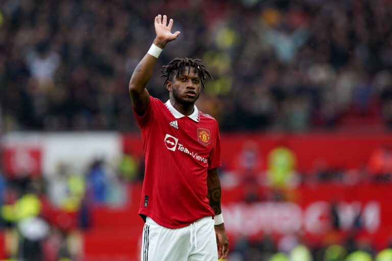 Manchester United footballer in a red kit raises his hand to the crowd during a match, stadium blurred in background.