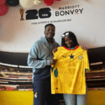Two people pose indoors with a yellow soccer jersey, in front of a FIFA World Cup 2026 Bonvoy backdrop with balloons nearby.