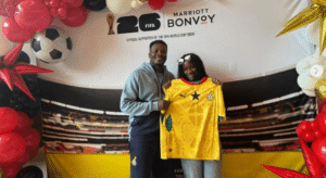 Two people pose indoors with a yellow soccer jersey, in front of a FIFA World Cup 2026 Bonvoy backdrop with balloons nearby.