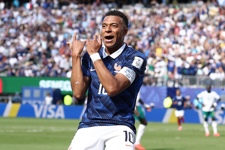 Soccer player in a blue kit celebrates a goal with arms raised as a packed crowd cheers in the stands.