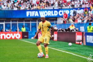 Soccer player in a yellow jersey (number 9) with a white headband, about to kick a ball on a bright green pitch at a FIFA World Cup match, stadium full of fans behind.