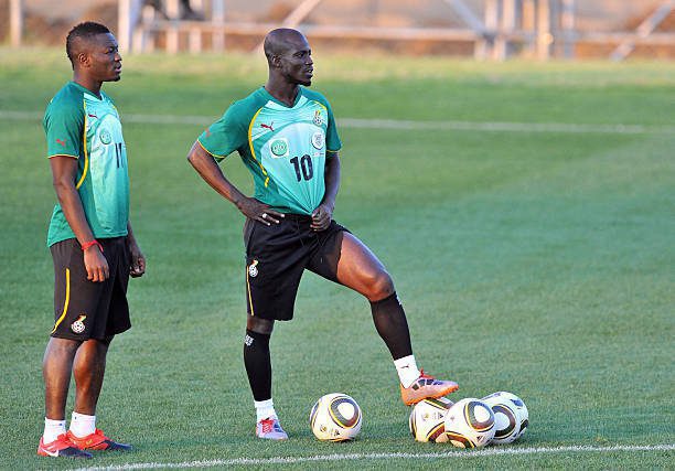 Two soccer players in green training kits stand on a grass field with several soccer balls at their feet.