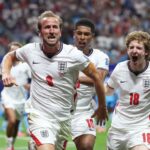 Three England players celebrate a goal on the pitch, wearing white jerseys with red numbers and an armband visible on one player.