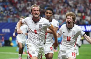 Three England players celebrate a goal on the pitch, wearing white jerseys with red numbers and an armband visible on one player.