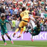 Soccer match moment: a yellow jersey player (27) leaps to head the ball while two green-jersey players (14 and 19) challenge from the ground in front of a crowded stadium.