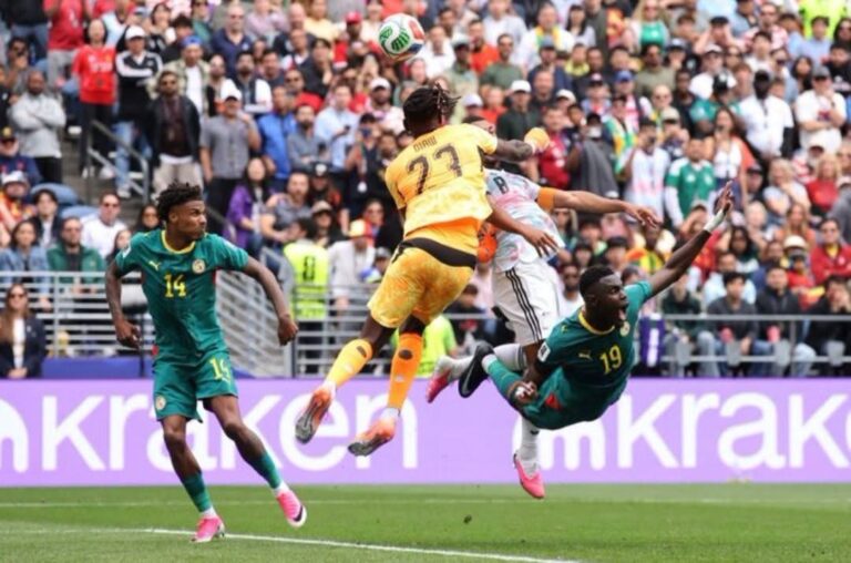 Soccer match moment: a yellow jersey player (27) leaps to head the ball while two green-jersey players (14 and 19) challenge from the ground in front of a crowded stadium.