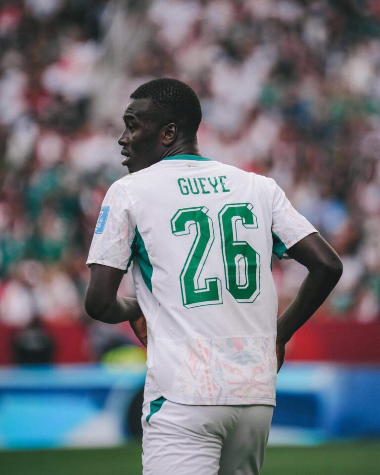 Soccer player seen from behind wearing a white jersey with green numbers and the name Gueye, number 26, on a crowded stadium backdrop