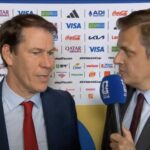 Two men in suits being interviewed on camera, one holding a blue microphone, with a backdrop covered in sponsor logos.