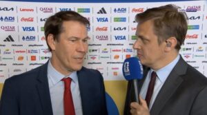 Two men in suits being interviewed on camera, one holding a blue microphone, with a backdrop covered in sponsor logos.