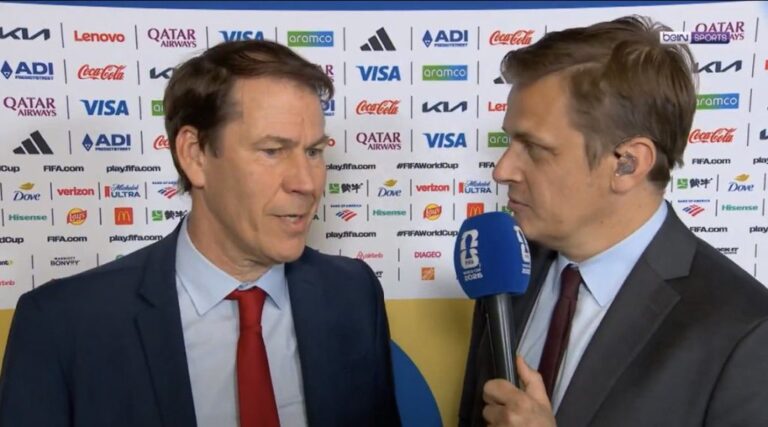 Two men in suits being interviewed on camera, one holding a blue microphone, with a backdrop covered in sponsor logos.