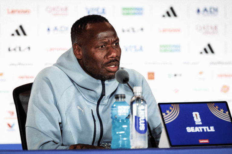 Athlete in a light blue jacket speaks into a microphone at a press conference; water bottles on the table and a Seattle banner on a laptop screen.