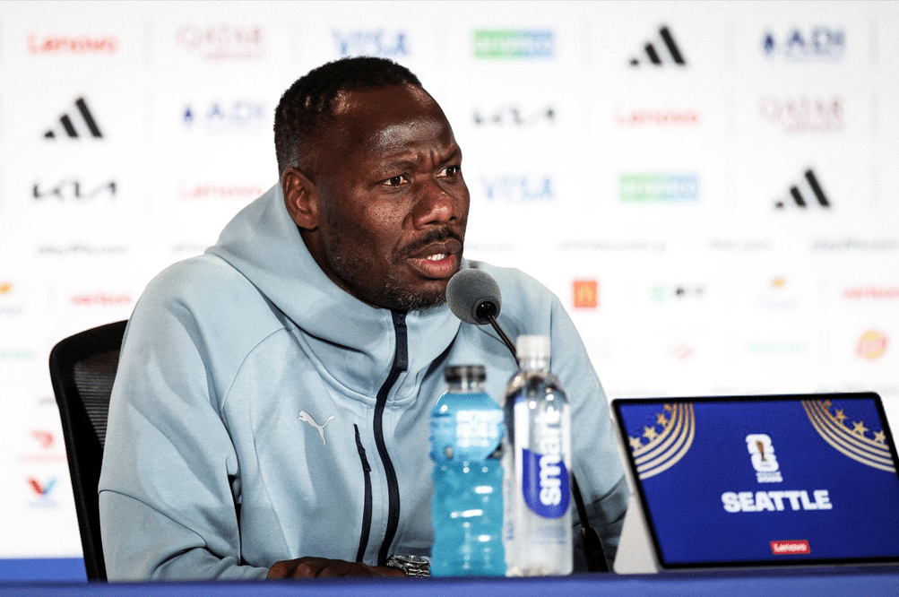 Athlete in a light blue jacket speaks into a microphone at a press conference; water bottles on the table and a Seattle banner on a laptop screen.