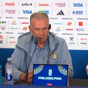 Man in a light blue jacket sits at a press conference desk wearing a headset, speaking into a microphone; backdrop shows sponsor logos, a water bottle on the left, and a laptop screen displaying 'PHILADELPHIA' with FIFA branding.