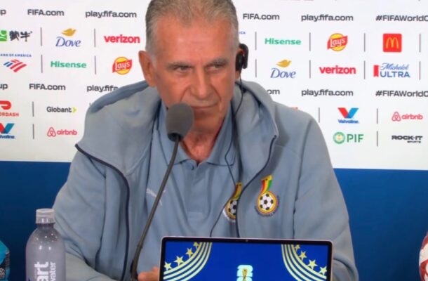 Man in a light blue jacket sits at a press conference desk wearing a headset, speaking into a microphone; backdrop shows sponsor logos, a water bottle on the left, and a laptop screen displaying 'PHILADELPHIA' with FIFA branding.