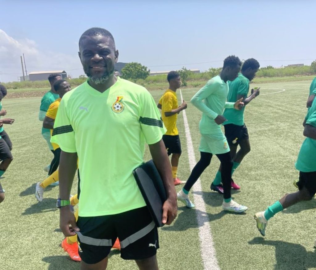 Coach or trainer standing on a sunny soccer field with a clipboard, surrounded by players in neon green jerseys warming up on artificial turf