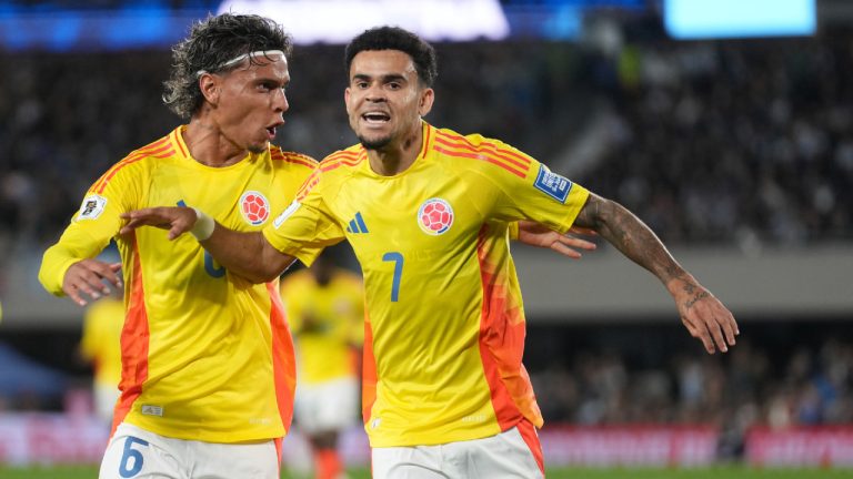 Two Colombian national team players in yellow jerseys celebrate on the soccer field, arms around each other after a goal or play.