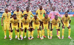 World soccer team posing for a pre-match photo on the field; 11 players in yellow uniforms with a pink-clad goalkeeper, stadium crowd in background.