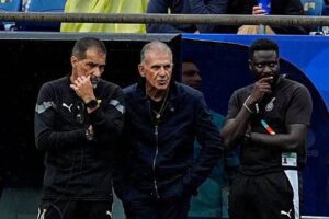 Three men on the sideline, two in dark tracksuits and a gray-haired coach in a dark jacket, watching the game with thoughtful expressions.