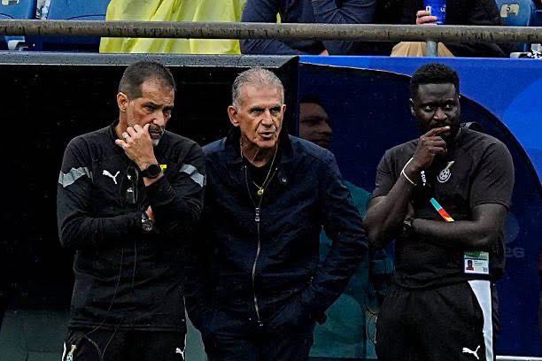 Three men on the sideline, two in dark tracksuits and a gray-haired coach in a dark jacket, watching the game with thoughtful expressions.