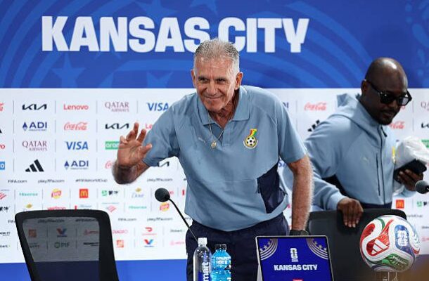IMG_1186 | Ghanaguardian.com Older man in a light blue polo waves at cameras during a Kansas City press conference, with a sponsor board, laptop, and water bottle on the table nearby.