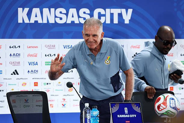 Older man in a light blue polo waves at a press conference with a 'Kansas City' banner in the background and sponsor logos on the wall behind him.