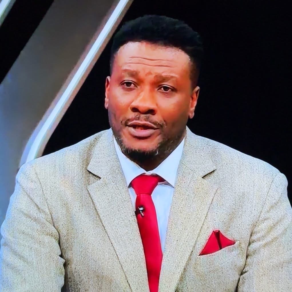 Man in a beige suit with a red tie speaking on a TV set.
