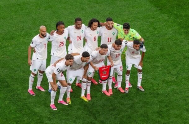 Soccer team in white jerseys huddles with arms around each other on a green grass field before the match.