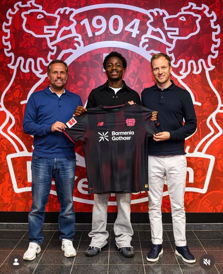 Three men pose indoors holding a dark red and black soccer jersey with sponsor logos (Barmenia Gothaer) in front of a red wall featuring a large white club crest and the number 1904.