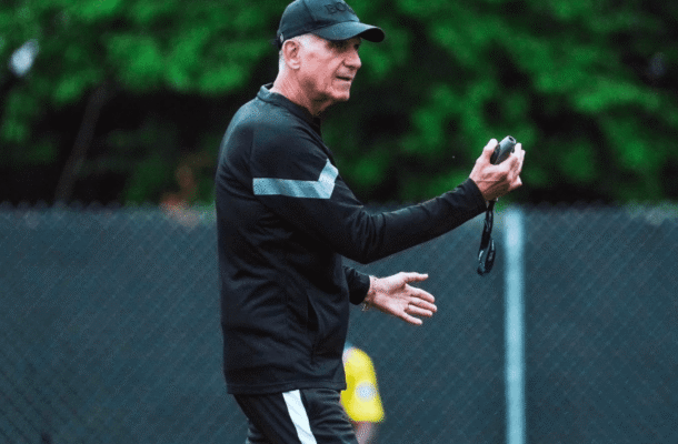 IMG_1191 | Ghanaguardian.com Male coach in a black tracksuit holds a stopwatch and speaks during outdoor practice on a chain-link fence field.