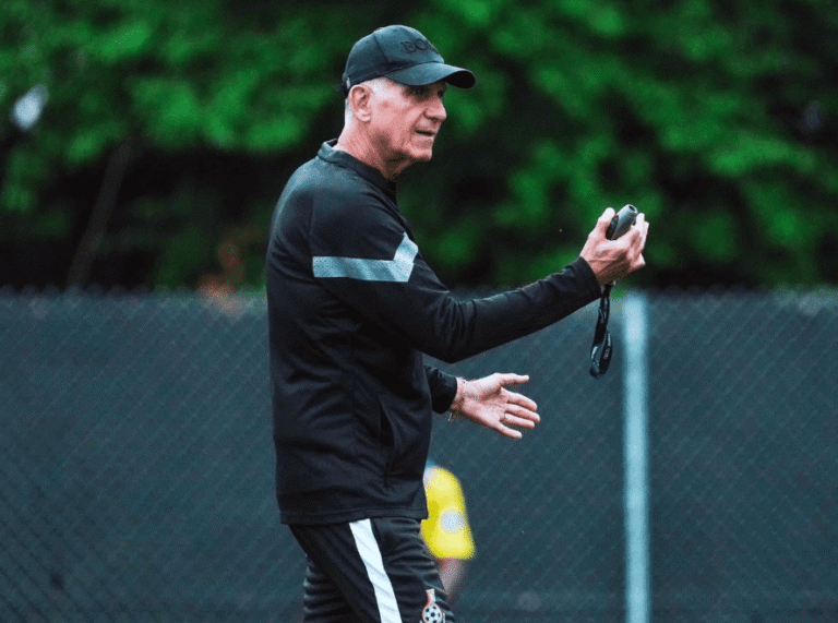 Male coach in a black tracksuit holds a stopwatch and speaks during outdoor practice on a chain-link fence field.