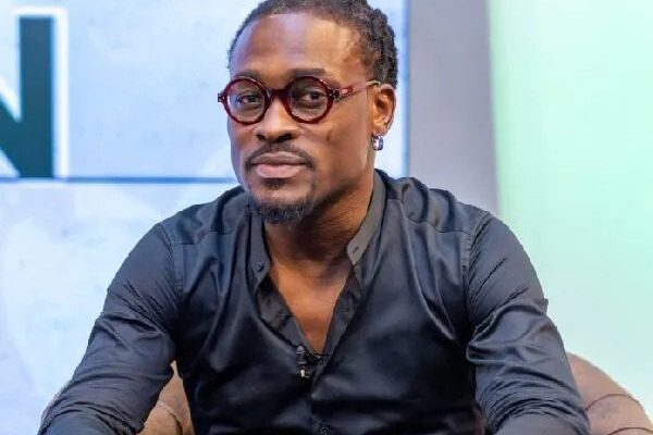 Portrait of a man with round red glasses and earrings, wearing a dark button-down shirt, seated in a studio setting.