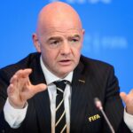 A bald man in a dark pinstripe suit speaks into a microphone, gesturing with his hands during a press conference, with a blue backdrop and a FIFA pin on his lapel.