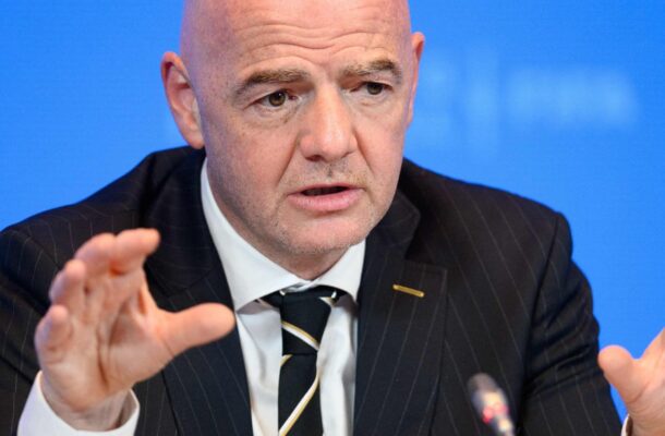 IMG_1199 | Ghanaguardian.com A bald man in a dark pinstripe suit speaks into a microphone, gesturing with his hands during a press conference, with a blue backdrop and a FIFA pin on his lapel.