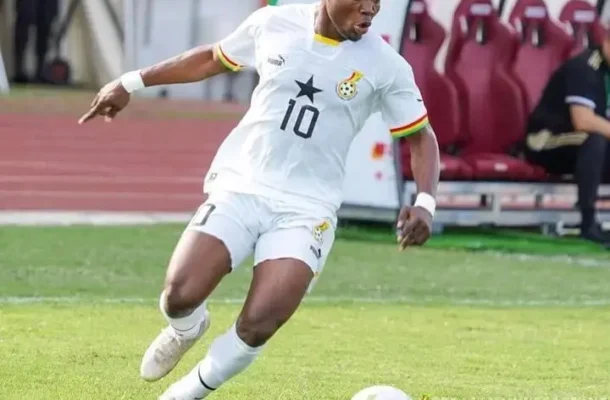 IMG_1200 | Ghanaguardian.com Soccer player in a white kit with number 10 dribbling a ball on a grassy field; substitutes bench visible in the background.