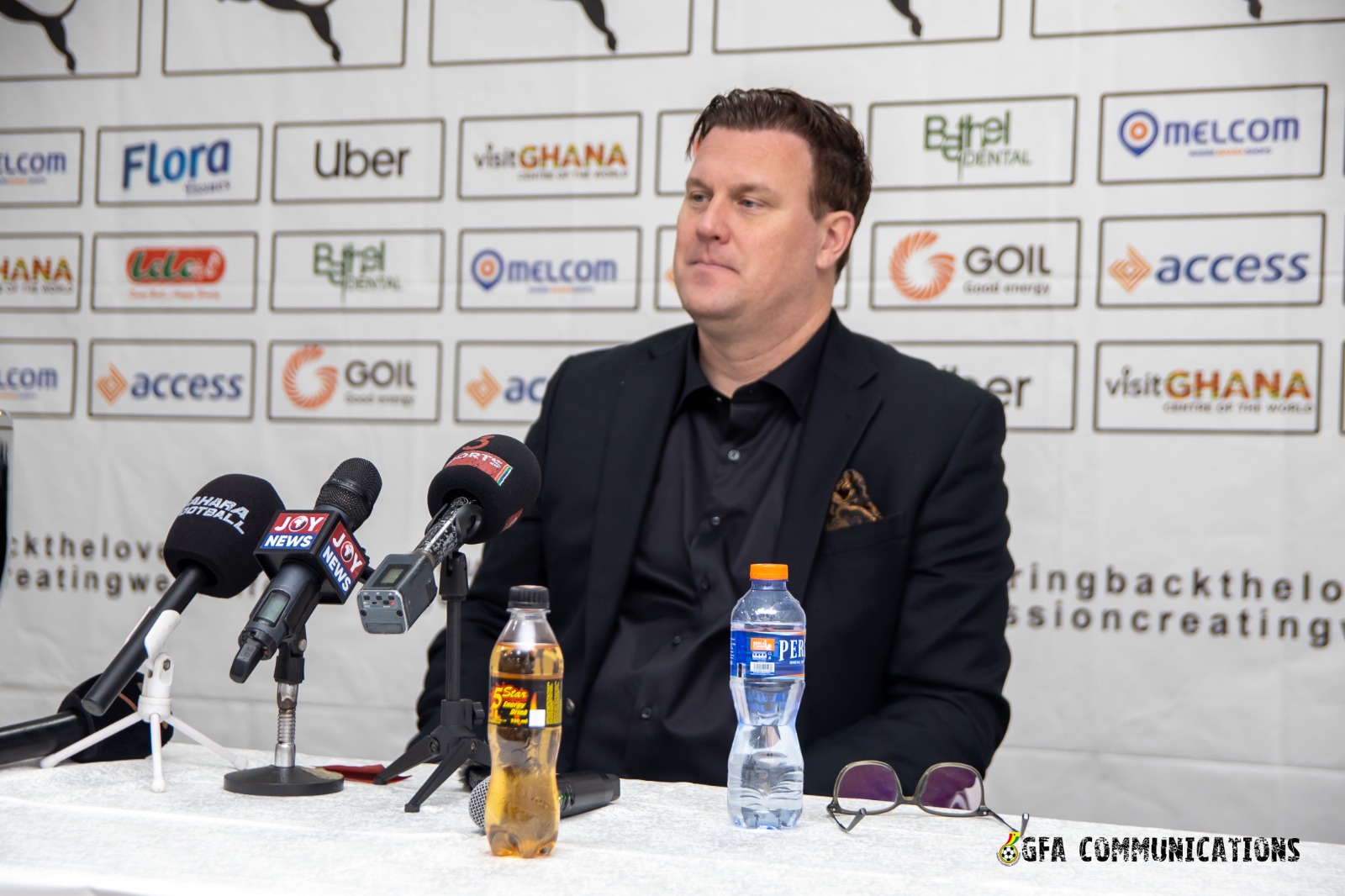 Man in a dark blazer seated at a press conference table with multiple microphones in front of him, sponsor logos on a backdrops behind him with water bottles on the table.