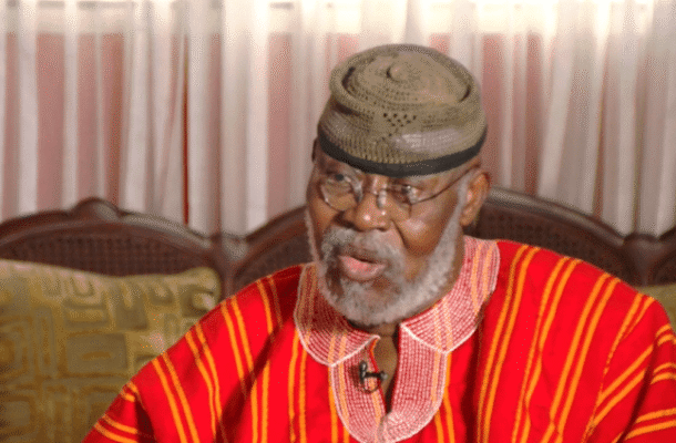 Older man with gray beard wearing a red striped traditional outfit and cap, seated indoors and speaking.] ,