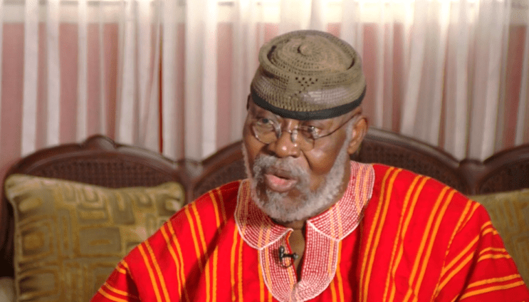 Older man with gray beard wearing a red striped traditional outfit and cap, seated indoors and speaking.] ,