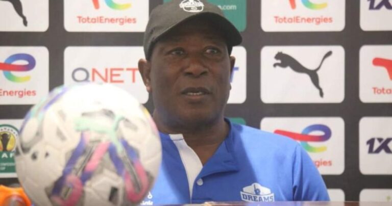 Man in a blue polo and cap speaks at a press conference with a soccer ball in the foreground and sponsor logos behind him.