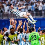 Argentina soccer players lift a teammate into the air in celebration on the field as fans cheer in the stands.