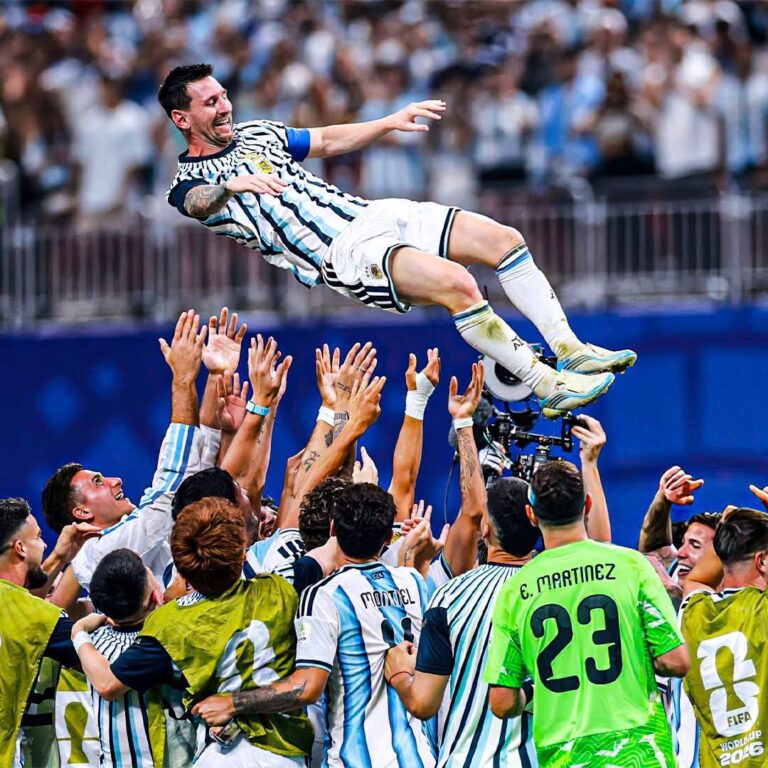 Argentina soccer players lift a teammate into the air in celebration on the field as fans cheer in the stands.