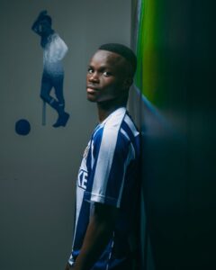 Young man in a blue-and-white striped soccer jersey leans against a wall, glancing at the camera in a dim hallway.
