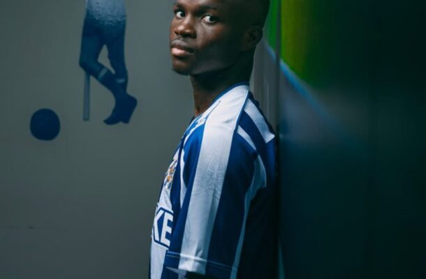 Young man in a blue-and-white striped soccer jersey leans against a wall, glancing at the camera in a dim hallway.